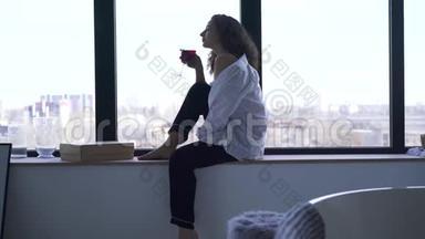 年轻女子坐在窗台上喝酒。 有思想的白人黑发女孩享受平静的侧面景色